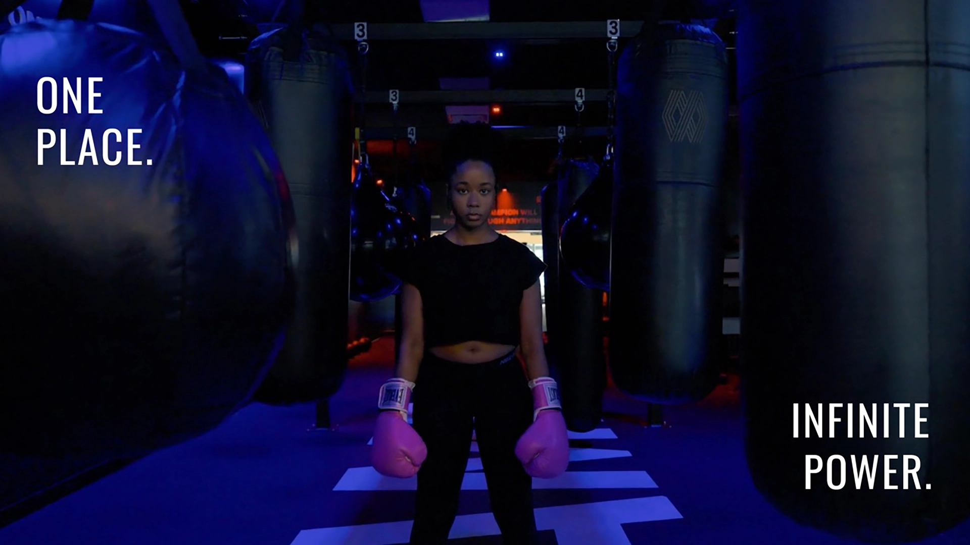 Woman stands in a boxing gym. Text reads "One place. infinite power."